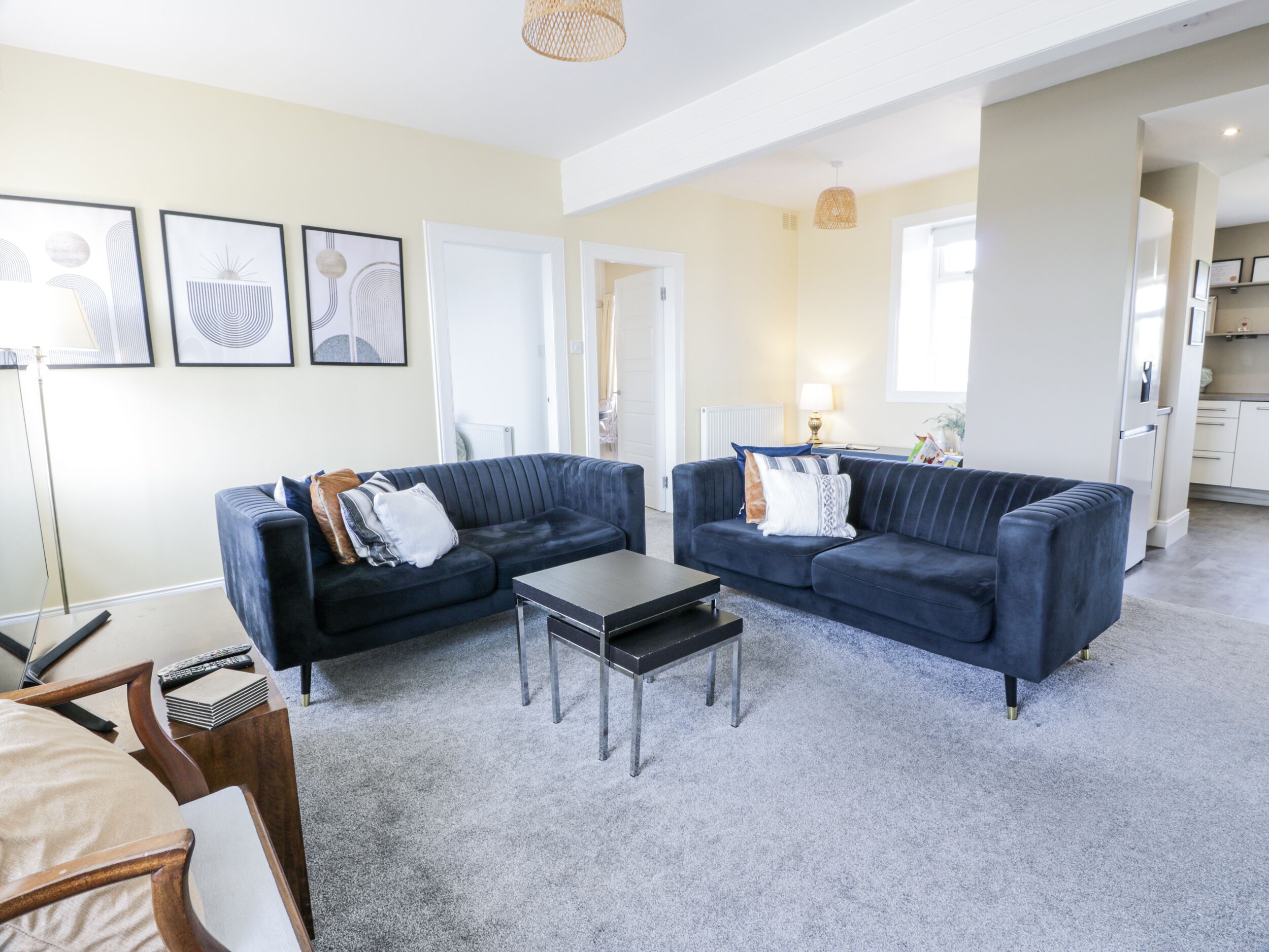 A living room with two dark blue sofas, a small coffee table, and a floor lamp. There are framed artworks on the wall and a kitchen area in the background.