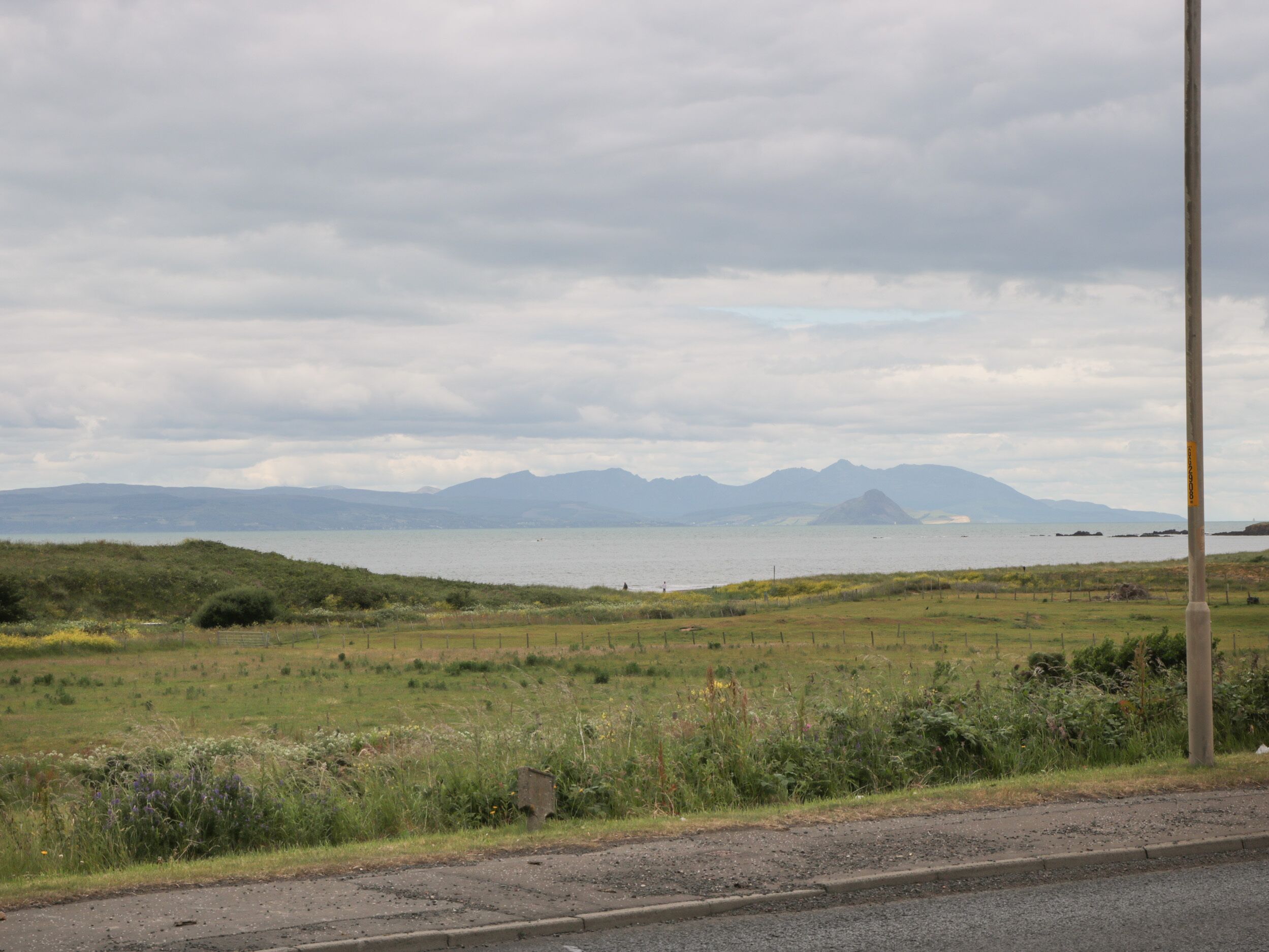 A coastal landscape with a road, grassy fields, and distant mountains.