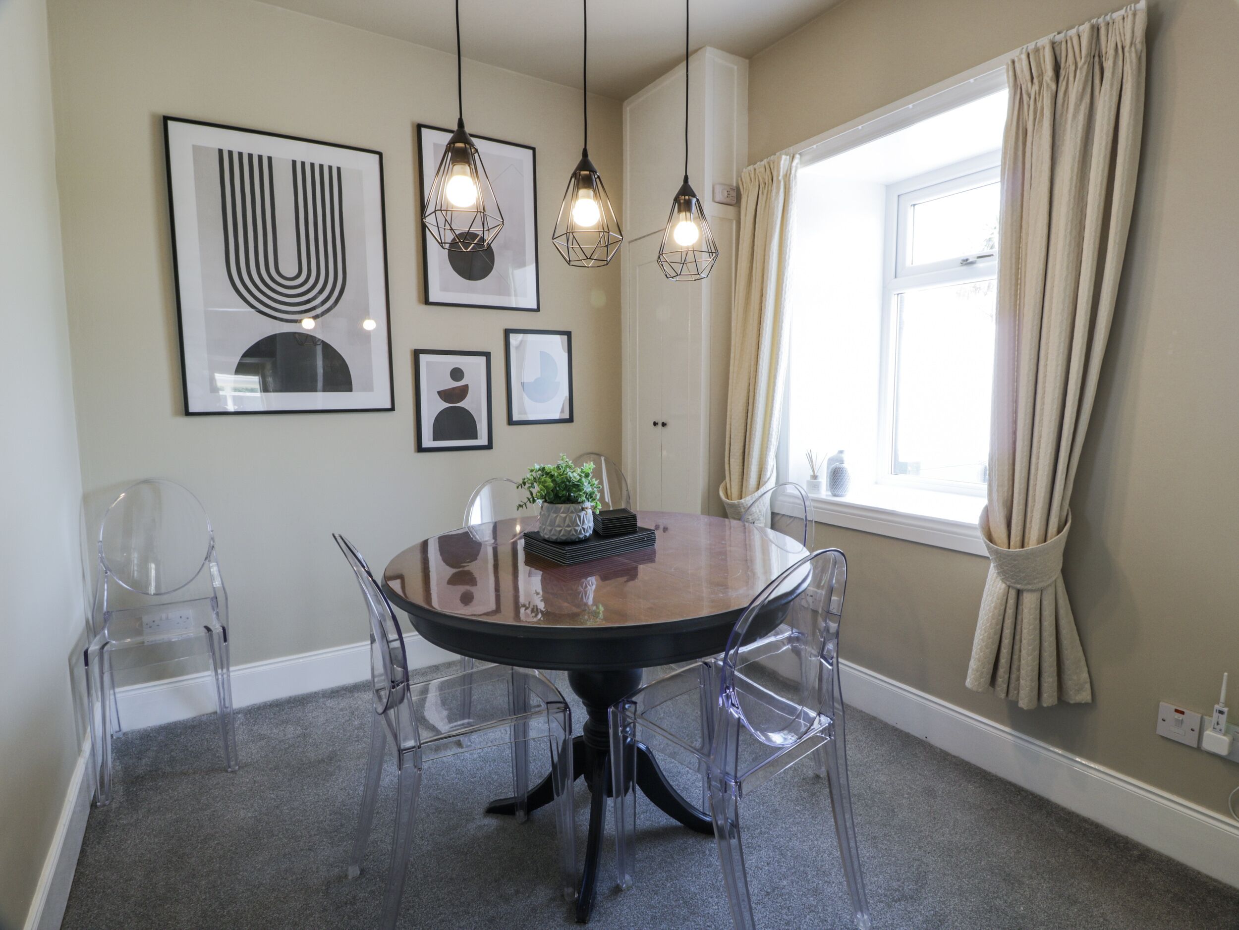 Un espace repas comprenant une table ronde, des chaises transparentes, des luminaires suspendus, des œuvres d’art encadrées et une fenêtre avec des rideaux.
