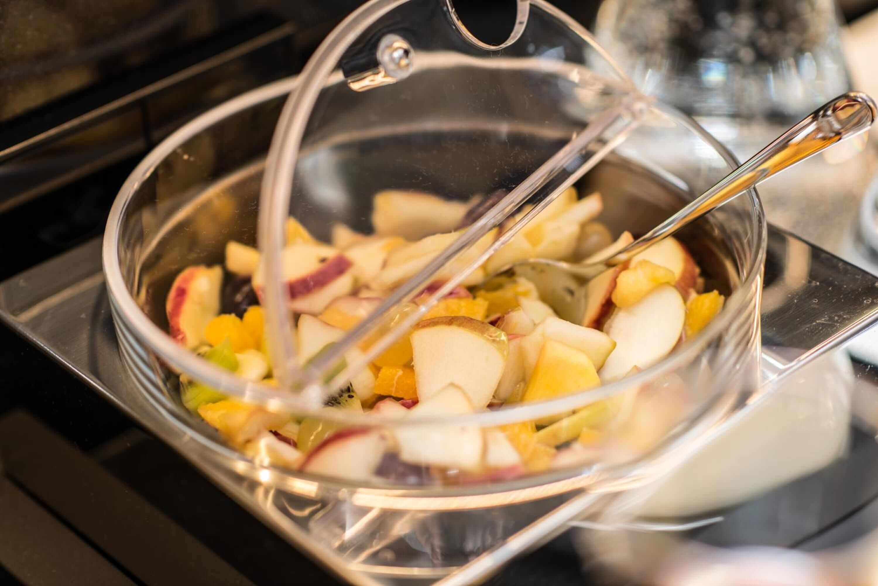 A glass bowl with a metal lid on a stove, containing chopped fruits and a spoon.