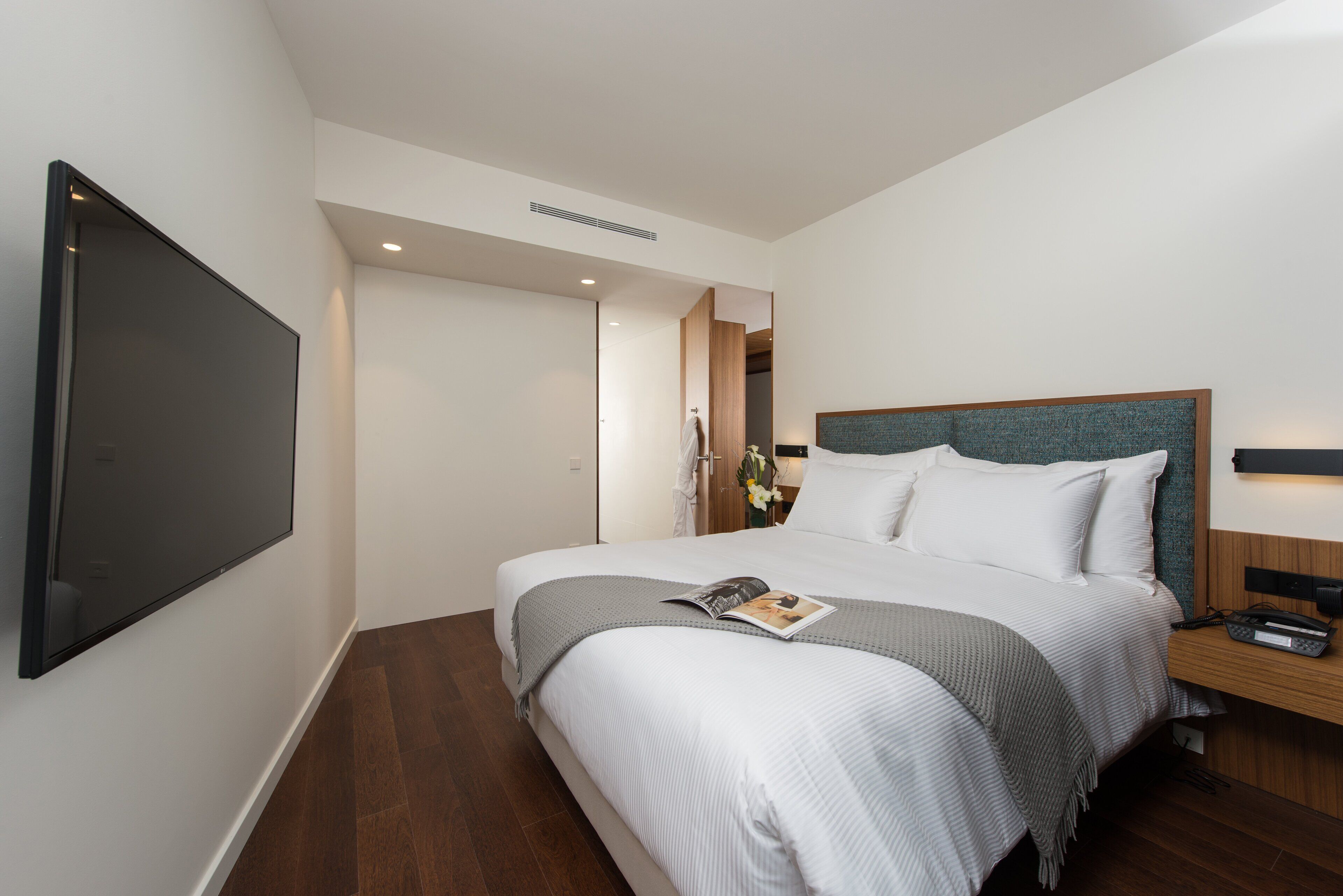 A modern hotel room with a large bed, a flat-screen TV mounted on the wall, and a wooden nightstand.