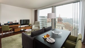 Habitación de hotel moderna con un amplio ventanal que ofrece vista a la ciudad, un comedor con mesa dispuesta para dos personas y una zona de estar con sofá y sillones.
