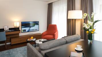 A modern hotel room with a sofa, a red armchair, a flat-screen TV, a coffee table with a cup and a tablet, and a vase with flowers.