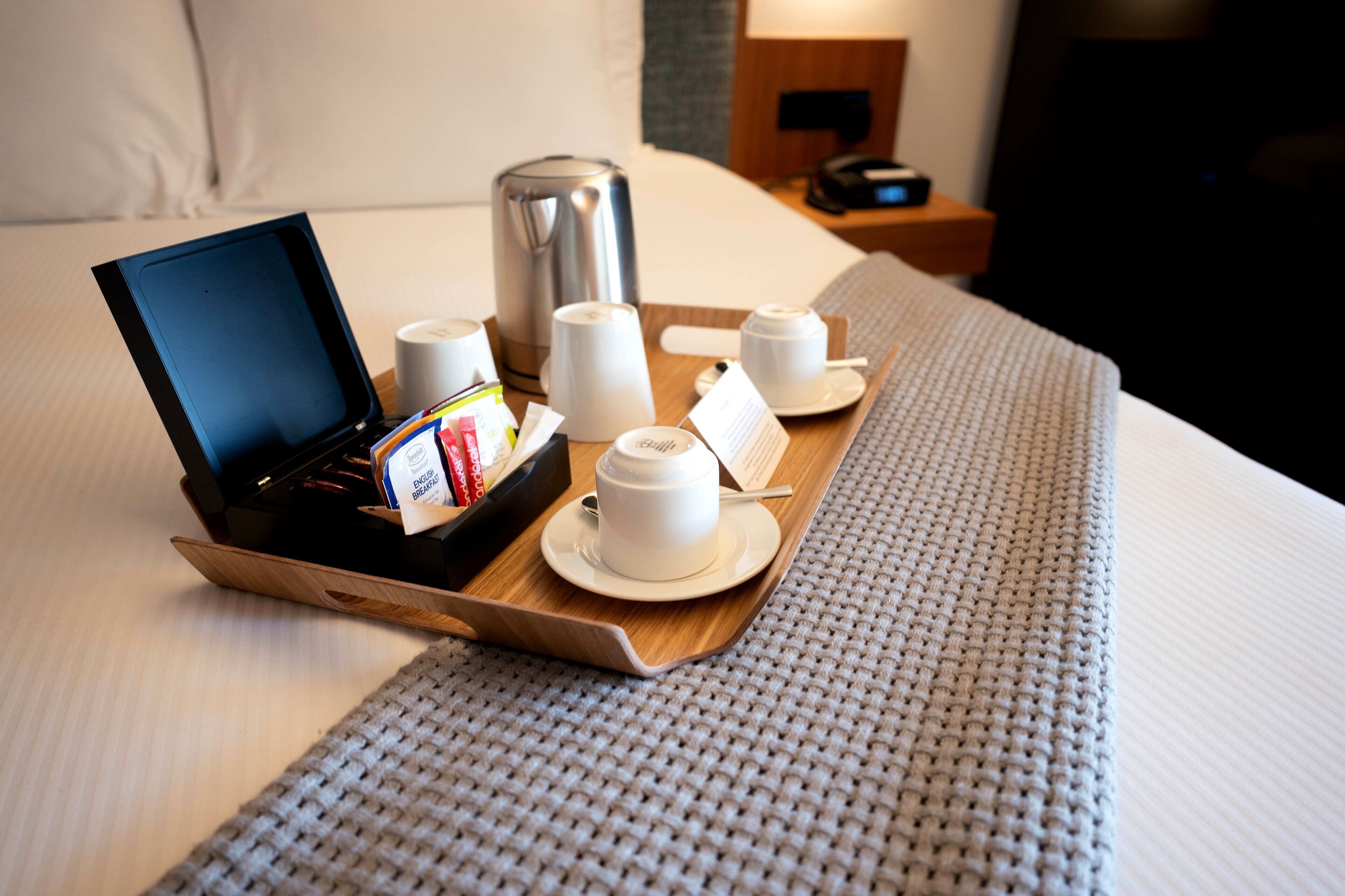 Un plateau de petit-déjeuner, joliment disposé, sur un lit, avec une machine à café, des tasses et une boîte de céréales.