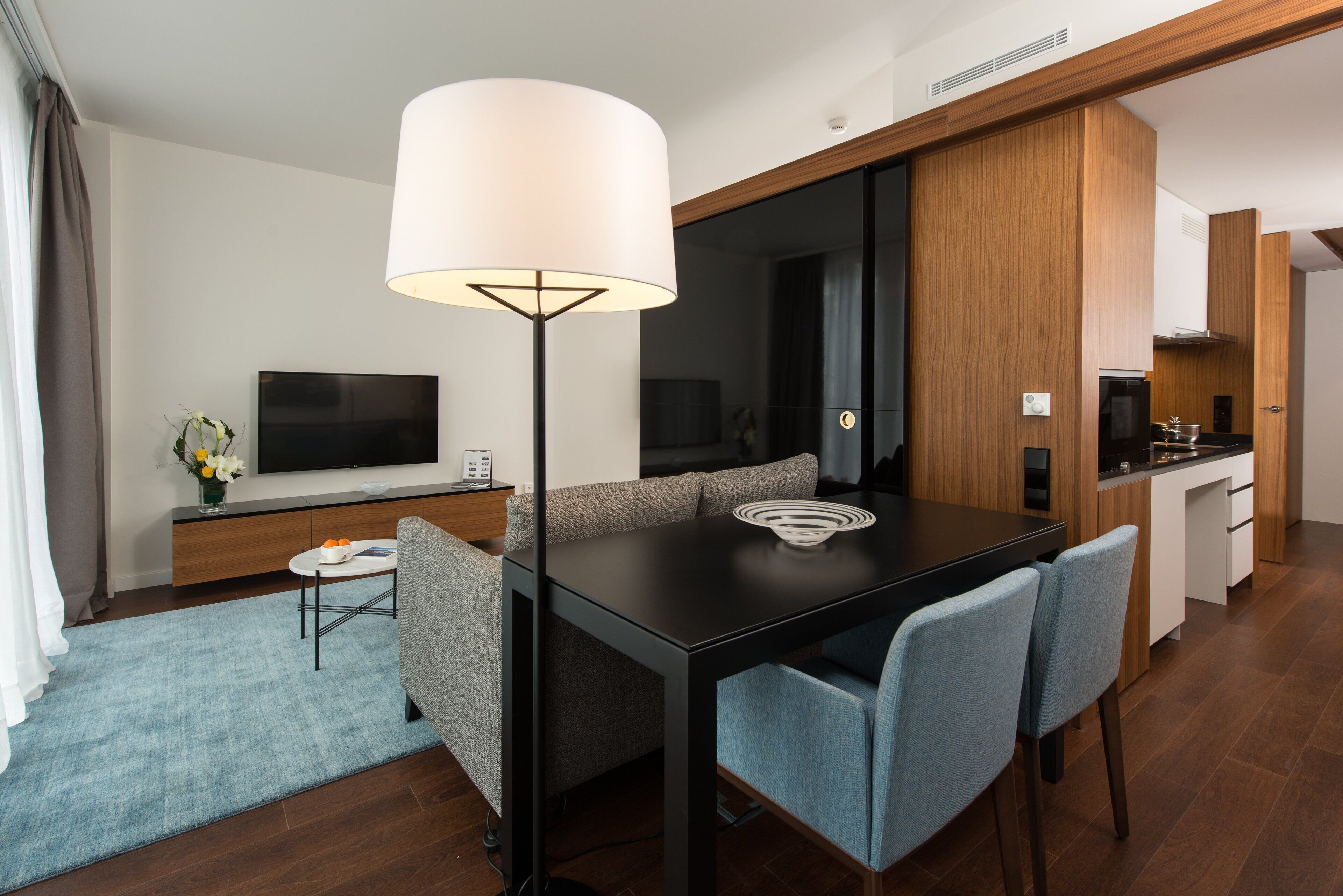 A modern hotel room with a dining area, a sofa, a TV, and a kitchenette.