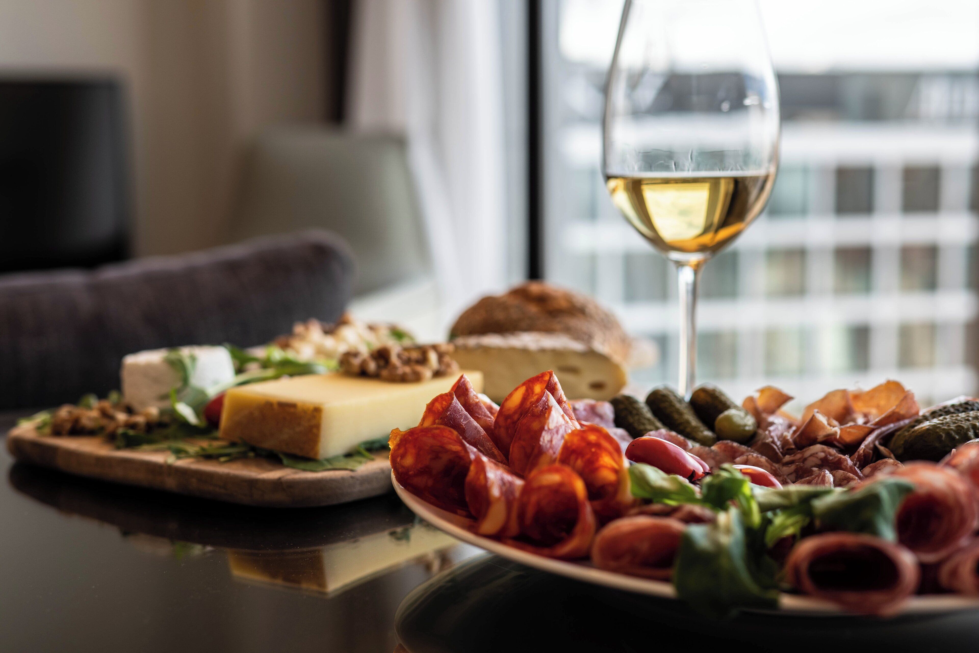 A table with a variety of cheeses, cured meats, and pickles, accompanied by a glass of white wine.