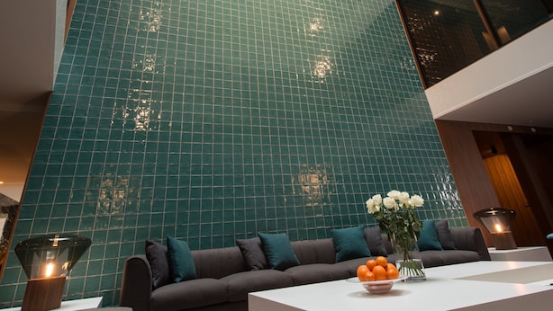 Un interior moderno con techo alto, pared de azulejos verdes, un sofá con cojines, una mesa de centro con un frutero y una mesita de noche con una lámpara.