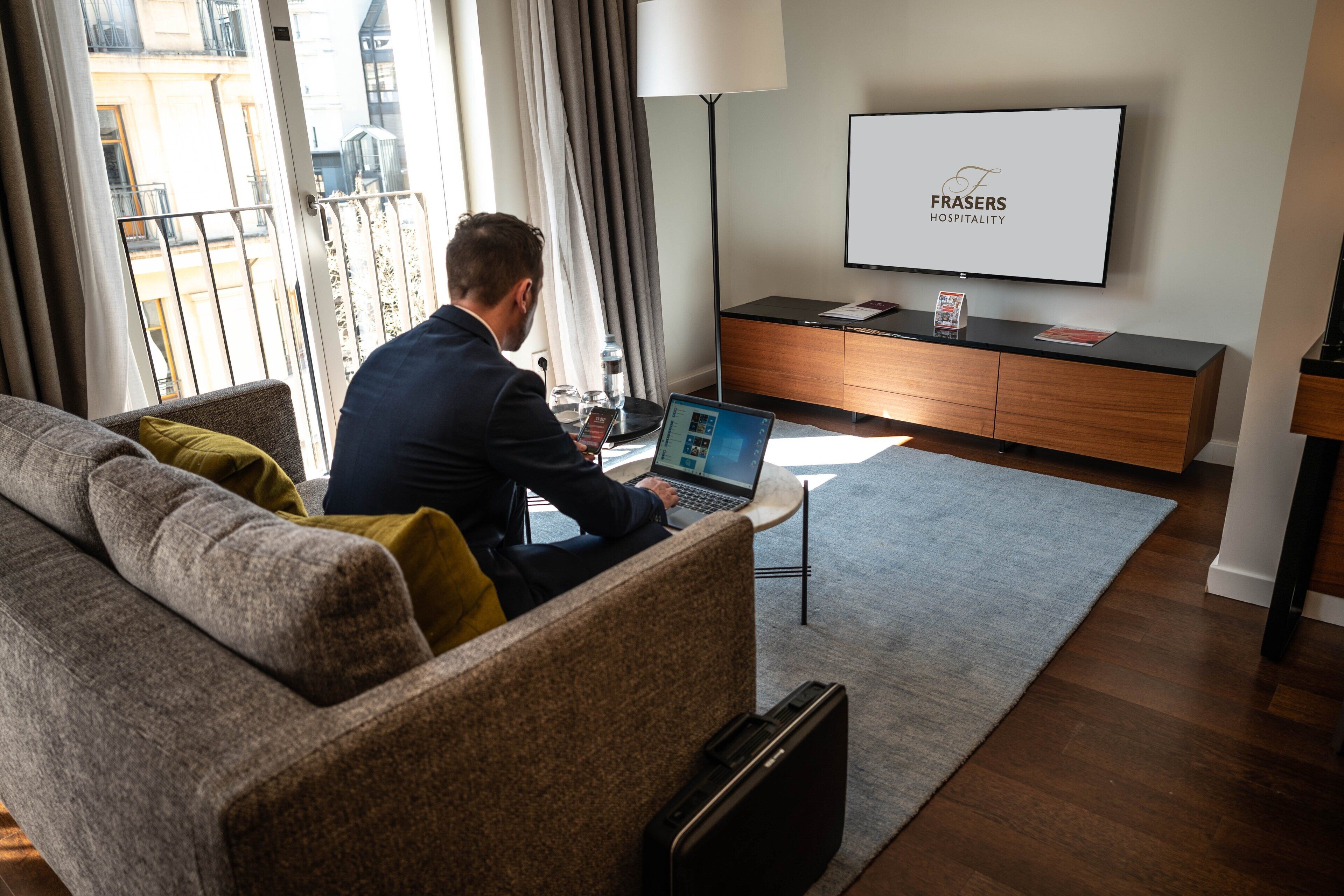 Un hombre trabaja en una computadora portátil en una habitación de hotel moderna con vista a la ciudad, un televisor de pantalla plana y una decoración minimalista.