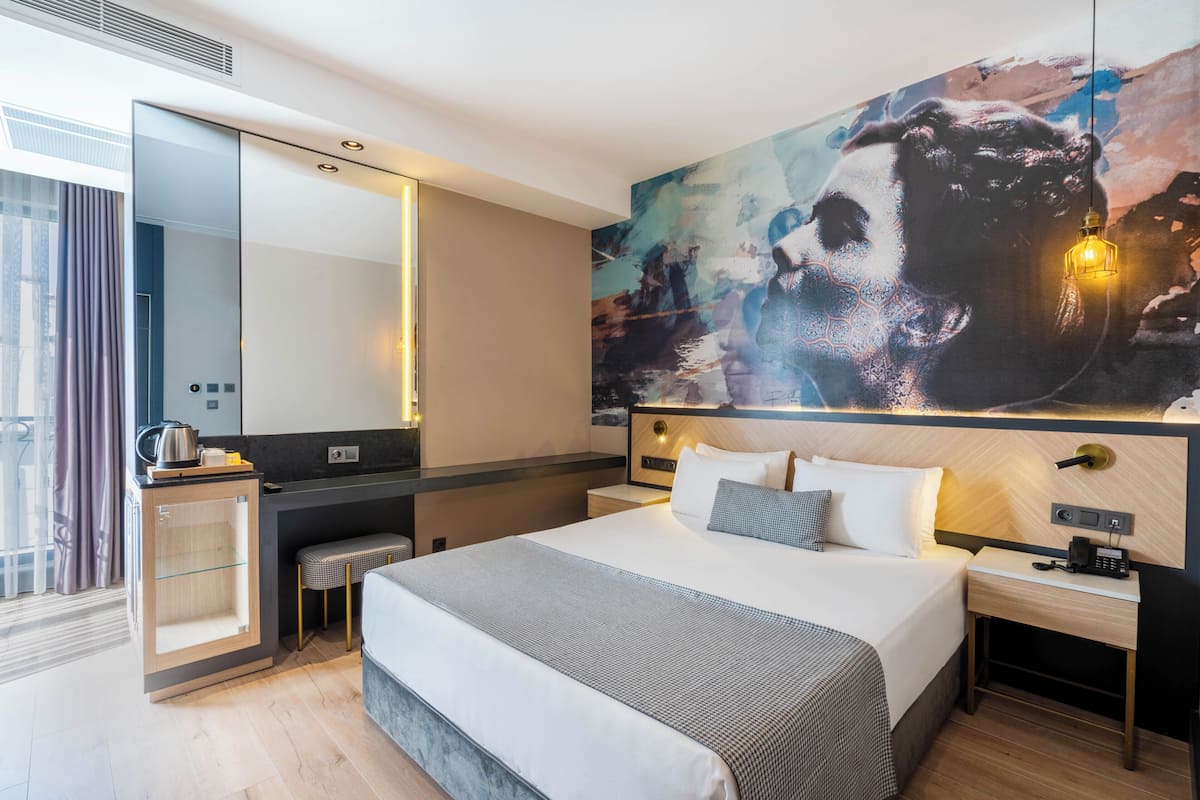 A modern hotel room with a large bed, a desk with a kettle, a mirror, and a large abstract painting on the wall.