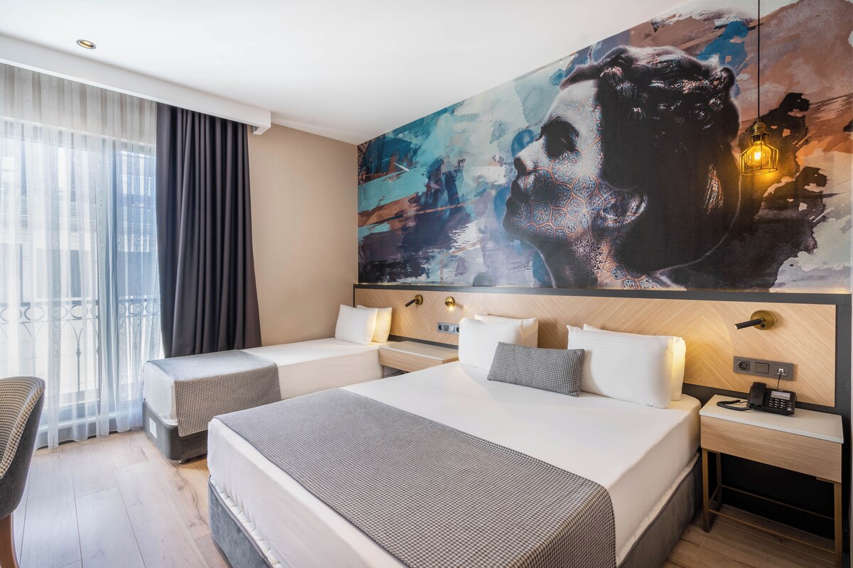 Una habitación de hotel con dos camas, un cuadro abstracto grande en la pared, una mesita de noche con una lámpara y vistas de la ciudad a través de cortinas translúcidas.