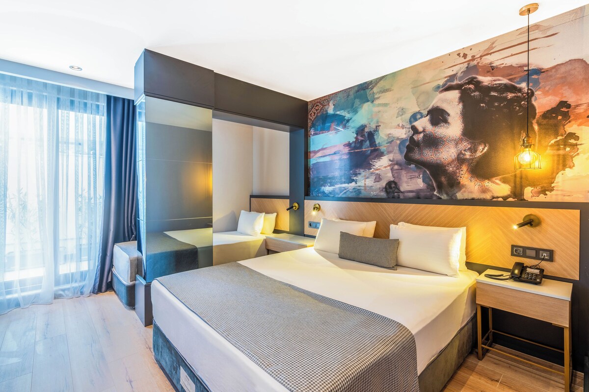 A modern hotel room with a large bed, bedside tables, a mirror, and a large abstract painting on the wall.