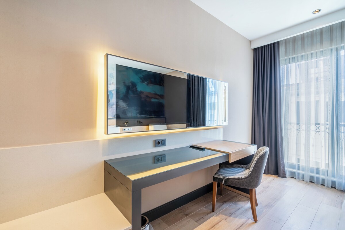A modern hotel room with a large flat-screen TV mounted on the wall, a sleek desk with a chair, and a large window with sheer curtains.
