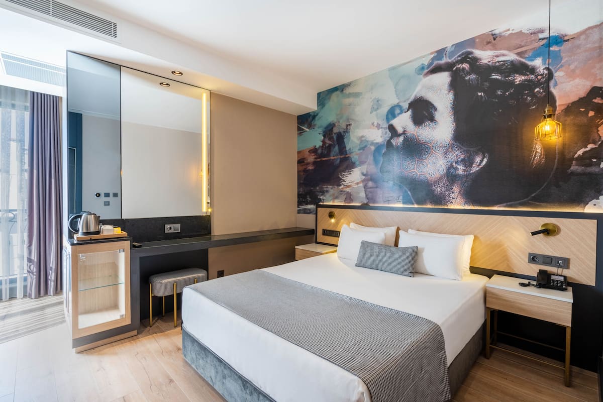A modern hotel room with a large bed, a desk with a kettle, a mirror, and a large abstract painting on the wall.