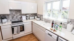 A modern kitchen with white cabinets, a stove, oven, and dishwasher. There is a window with blinds, a countertop with various items, and a tiled backsplash. - 3 Rothbury Place (Lytham St. Anne's)