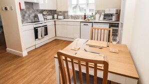 A modern kitchen with white cabinetry, a wooden dining table, and a window with blinds. - 3 Rothbury Place (Lytham St. Anne's)