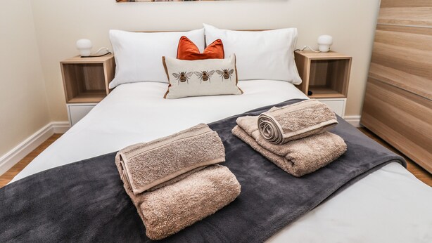 A neatly made bed with a beach scene painting above it, two nightstands with lamps, and a wooden headboard. - 3 Rothbury Place (Lytham St. Anne's)
