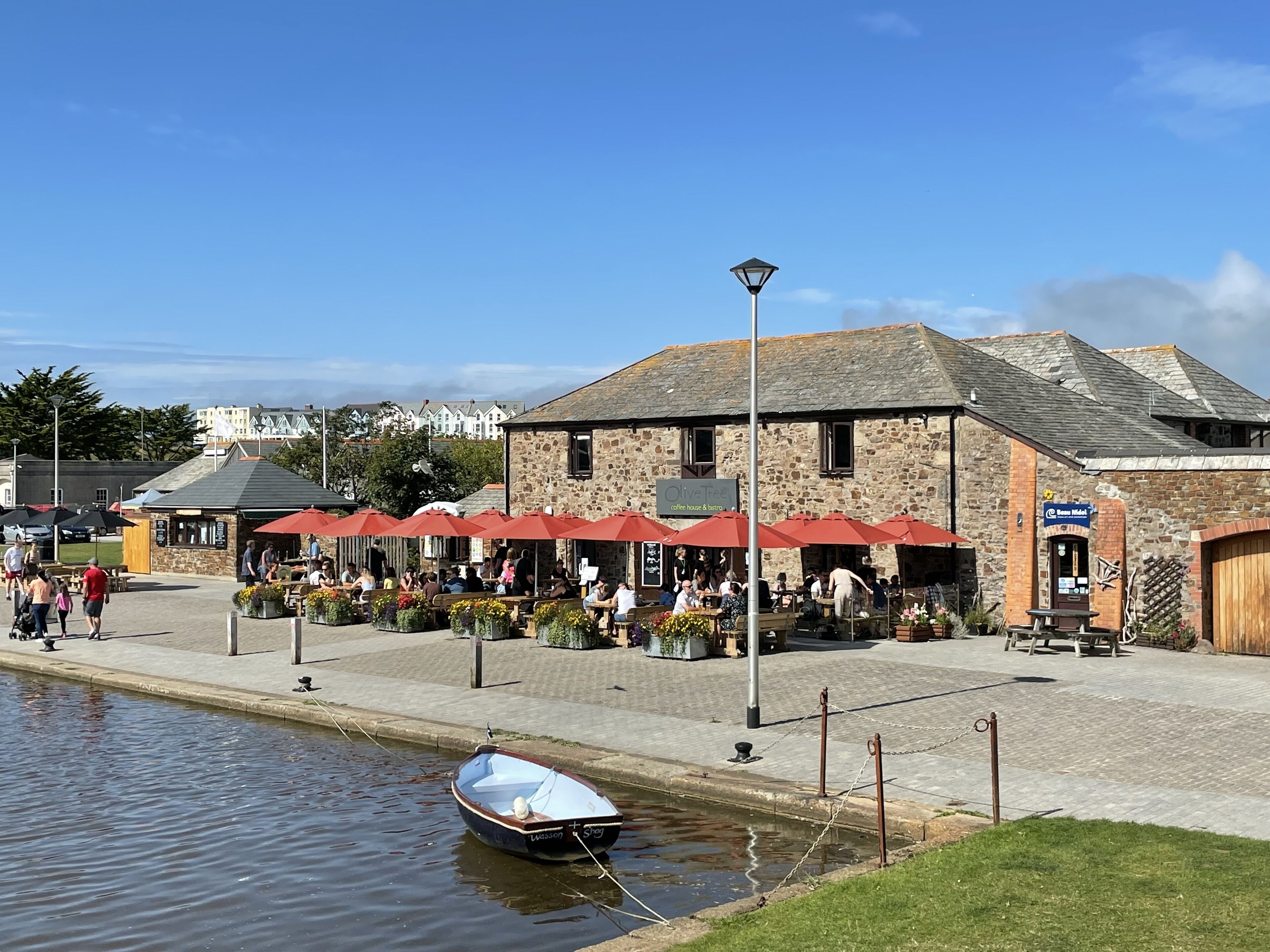 A riverside café with outdoor seating, a stone building, and a small boat docked by the water.