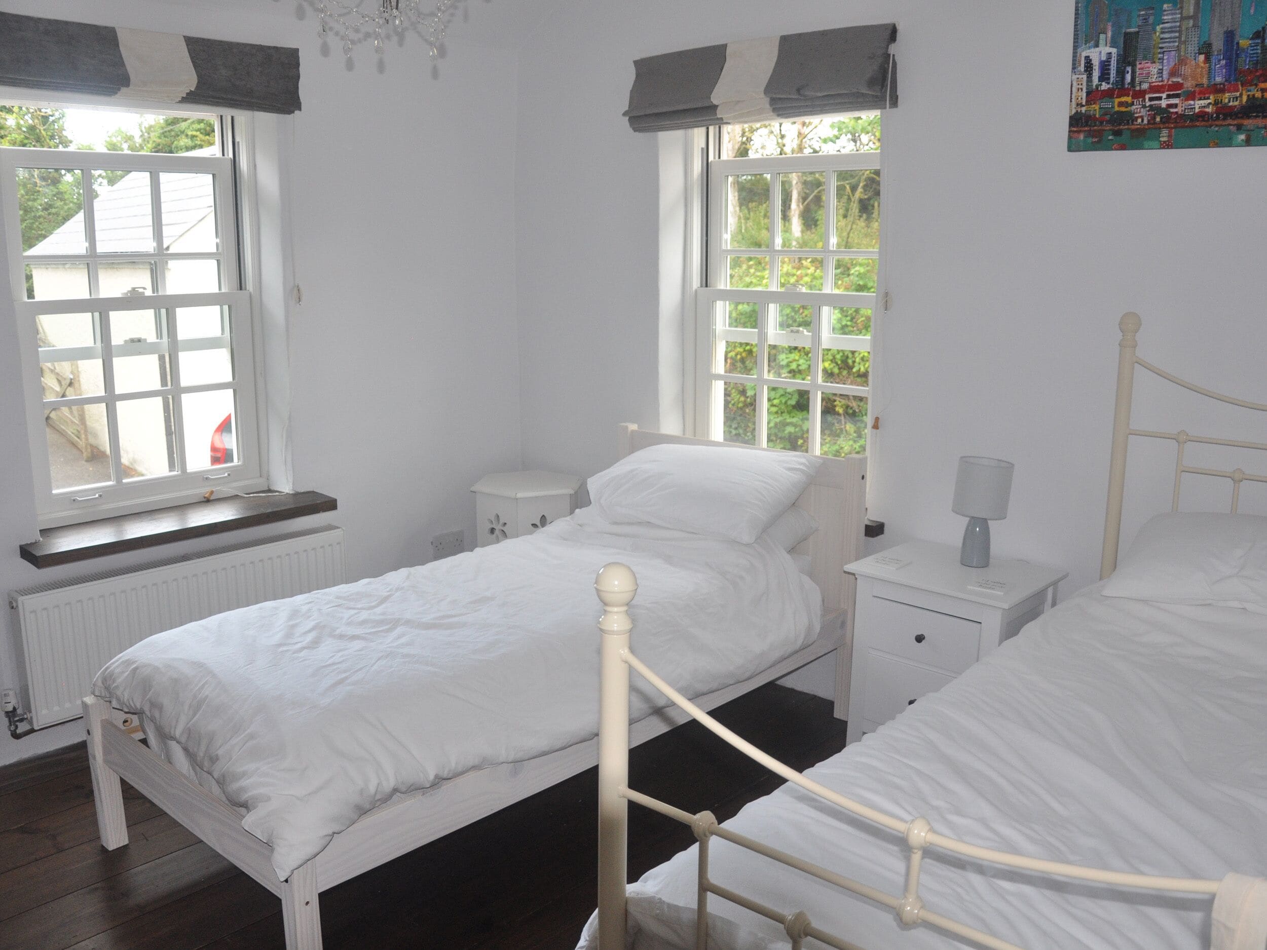 Un dormitorio con dos camas individuales, una ventana con vista a la vegetación, un radiador y una mesita de noche pequeña con lámpara.