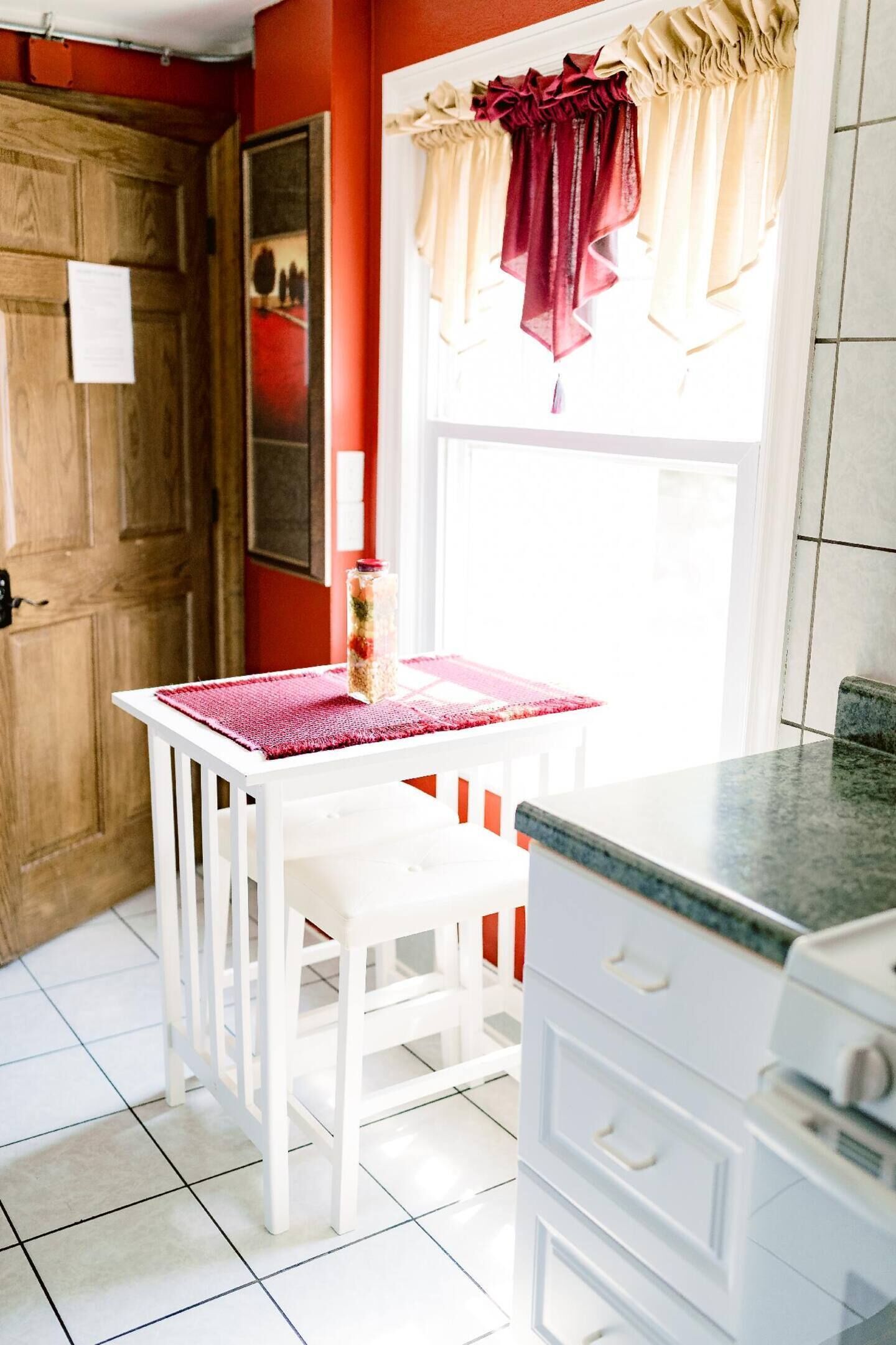 Una cocina con armarios blancos, una mesa blanca con un mantel individual rojo y una ventana con cortinas rojas.