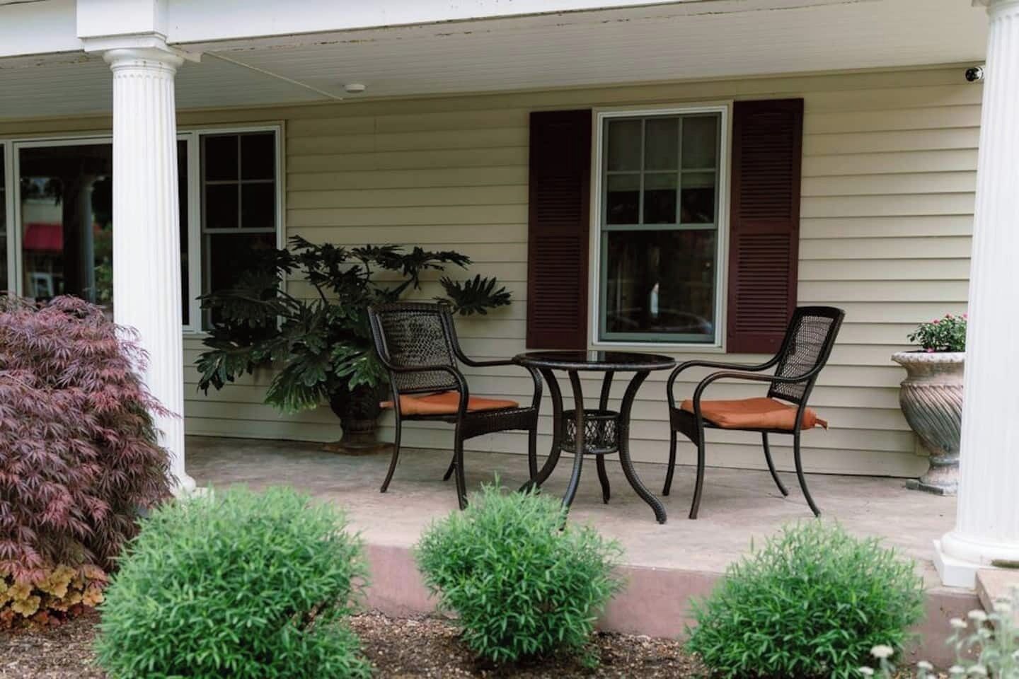 A porch with two chairs and a table, a large potted plant, and trimmed bushes.