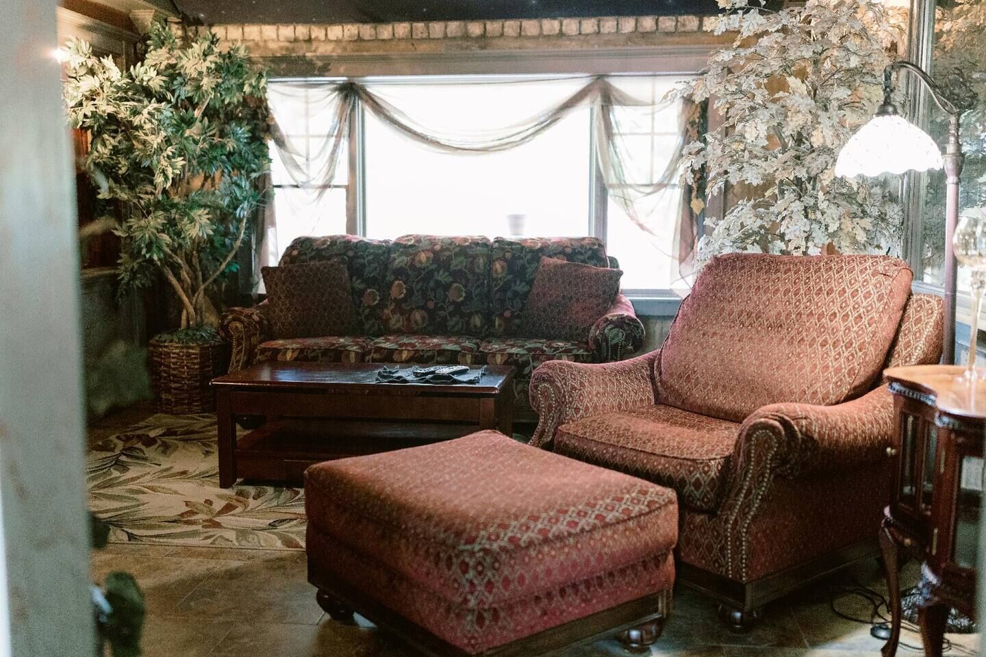 Un salón acogedor con un sofá estampado, un sillón a juego y un reposapiés coordinado. Hay una planta grande en maceta y una lámpara sobre una mesita auxiliar.