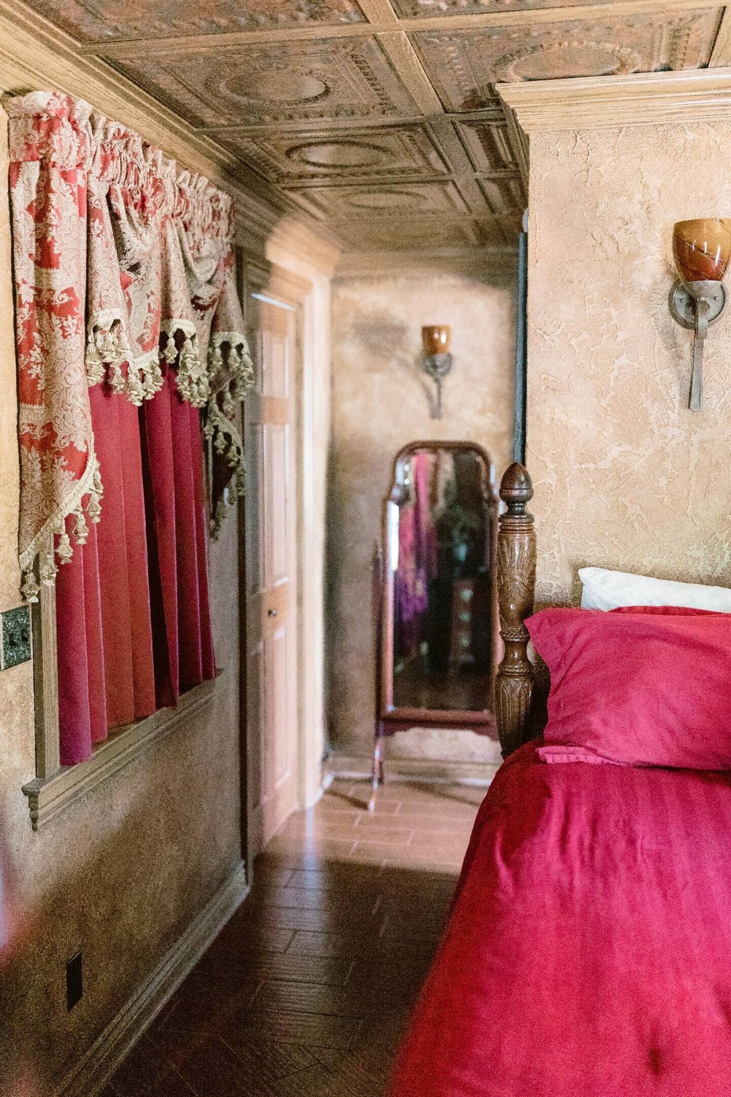 Un couloir étroit avec une porte en bois, un miroir et un lit recouvert d’une literie rouge.