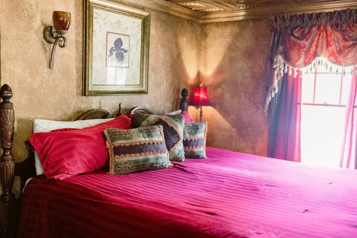 A bed with a red bedspread and decorative pillows, a framed picture on the wall, and a red lamp on a bedside table.