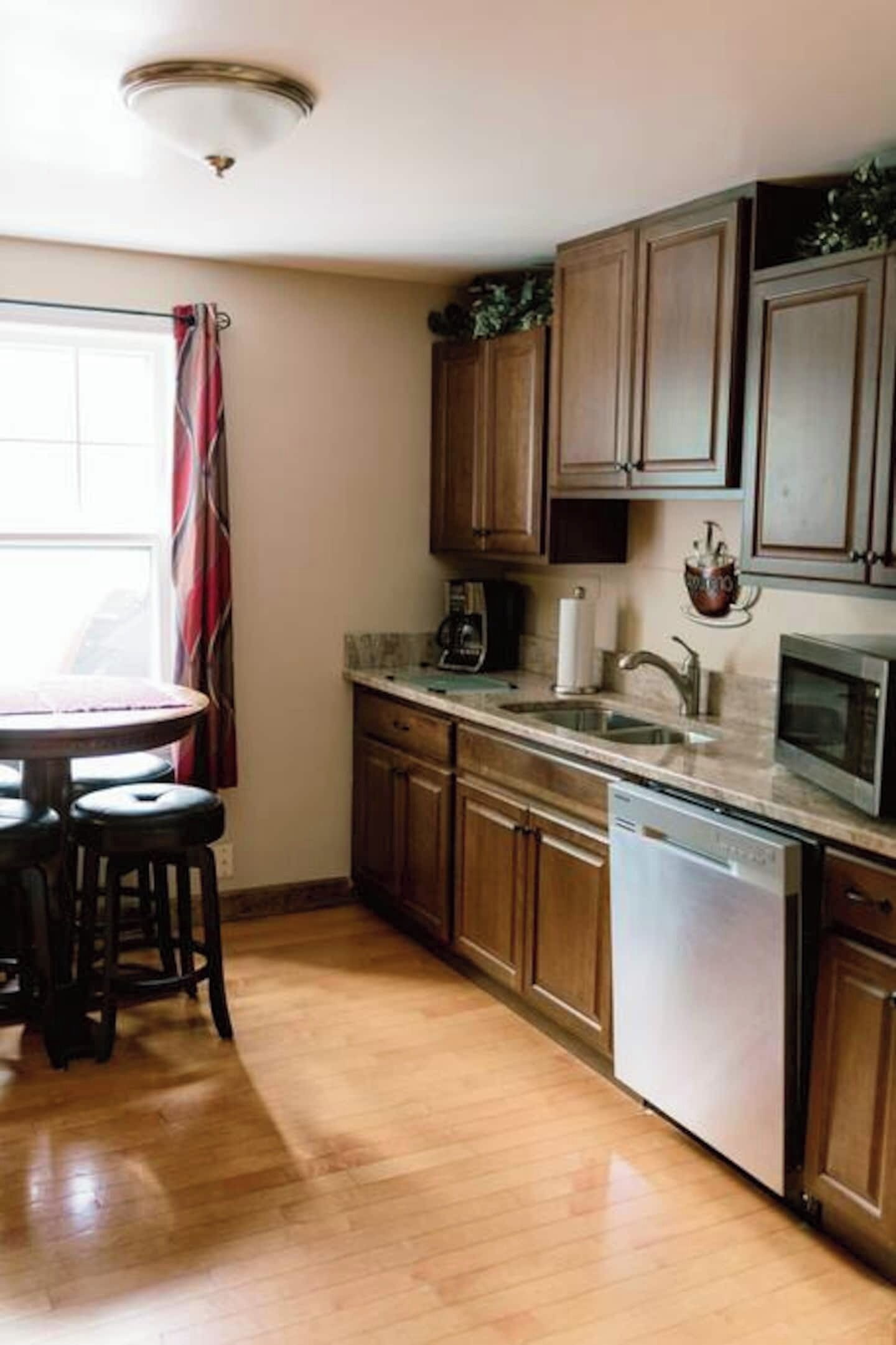 A kitchen with wooden cabinets, a stainless steel dishwasher, a microwave, and a dining area with a round table and stools.