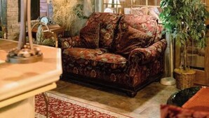 Une pièce chaleureuse avec un canapé à motifs, une lampe, une bibliothèque et une grande fenêtre donnant sur des immeubles.