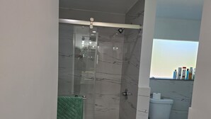 Moderni kylpyhuone, jossa on lasiseinällä varustettu suihku, wc ja avoin hylly, jossa on hygieniatuotteita.