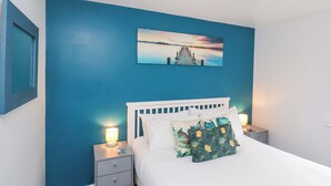 A bedroom with a white bed, blue walls, a nightstand with a lamp, and a picture of a sunset over water. - Dabbling Ducks (Whitby)