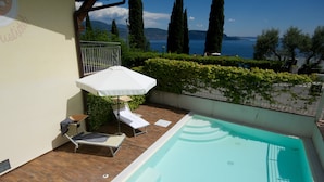 Una piscina con sillones de descanso y una sombrilla, rodeada de follaje y vista al mar.
