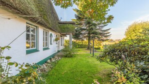 A traditional thatched-roof house with a white exterior and green trim, surrounded by a well-maintained garden with various plants and a clear sky.