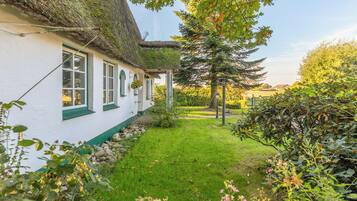 A traditional thatched-roof house with a white exterior and green trim, surrounded by a well-maintained garden with various plants and a clear sky.