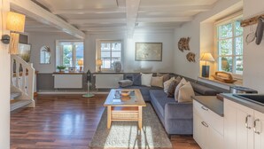 A modern living room with a wooden floor, a grey sofa, a coffee table, and a kitchen area with white cabinets and a window.