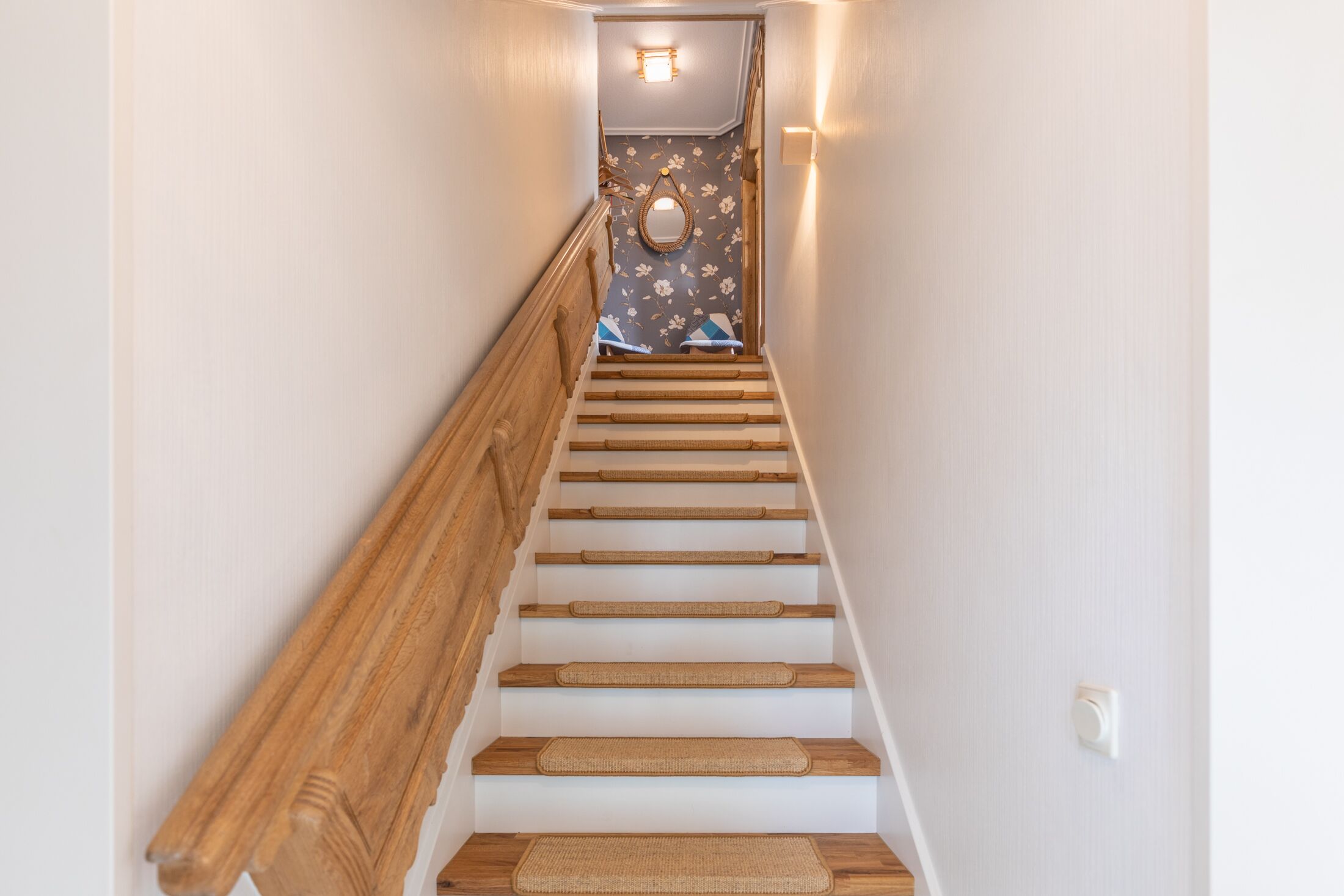 Un escalier en bois avec une rampe, menant à une pièce dont les murs sont recouverts d’un papier peint à motifs floraux et qui contient un miroir.