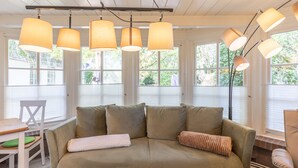 Una habitación acogedora con un sofá verde, una mesa de madera y una silla. Amplias ventanas que dejan entrar luz natural y ofrecen vistas al exterior.