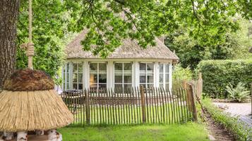 A thatched-roof gazebo with white-framed windows, surrounded by a wooden fence and lush greenery.