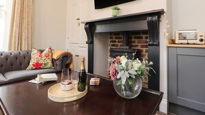 A living room with a fireplace, a flat-screen TV mounted above it, a sofa with floral cushions, and a coffee table with a vase of flowers and glasses. - No 19, Haworth (Keighley)