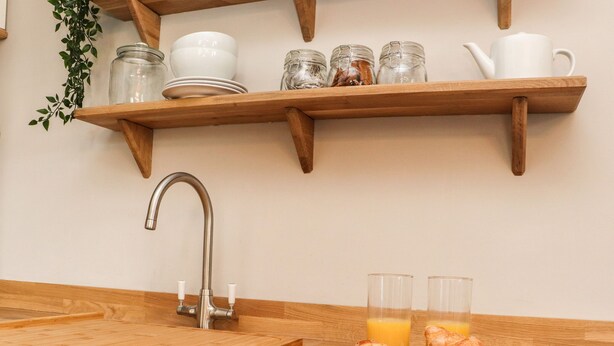 A kitchen with wooden shelves holding dishes, jars, and a teapot. There is a sink with a faucet, a wooden cutting board, and a plate with croissants and strawberries. - No 19, Haworth (Keighley)