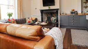 A living room with a leather sofa, a fireplace, a flat-screen TV, and a kitchen area with a microwave and toaster. - No 19, Haworth (Keighley)