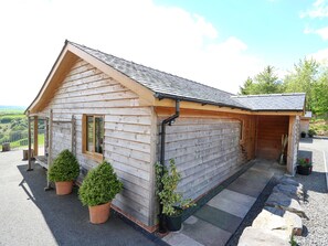A wooden cabin with a sloped roof, a porch with potted plants, and a stone pathway leading to the entrance. - The Larches (Llanidloes)