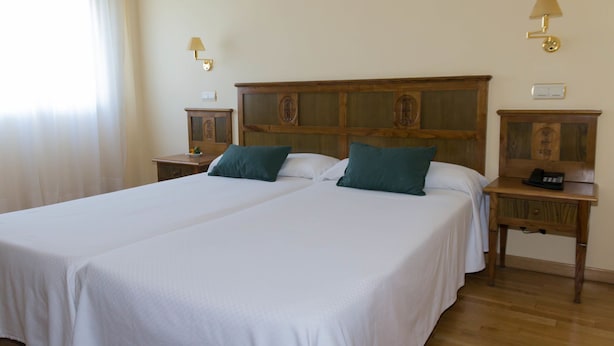 A double bed with two pillows, a wooden headboard, bedside tables with lamps, and a small clock. - Hotel ELE Puerta de Monfrague (Malpartida de Plasencia)