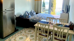 A compact hotel room with a bed, a small table, a chair, a refrigerator, and a window with a view of a balcony and greenery.