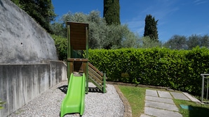 A playground with a slide and a climbing structure, surrounded by a hedge and a concrete wall.