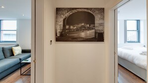 Et moderne hotelværelse med et stort vindue, en seng med hvide sengetøj, en blå sofa med pude med mønster og et vægmaleri, der viser et bybillede.
