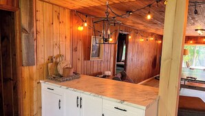 Un interior de madera con una zona de cocina que incluye encimera de mármol, armarios blancos y luces colgantes.