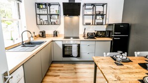 A compact kitchen with a sink, stove, oven, and black refrigerator. There are shelves above the counter holding dishes and glasses. - Victoria House (Holmfirth)