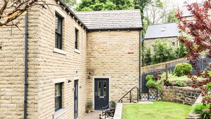 A stone building with a black door, a small garden with a bench, and a fence. - Victoria House (Holmfirth)