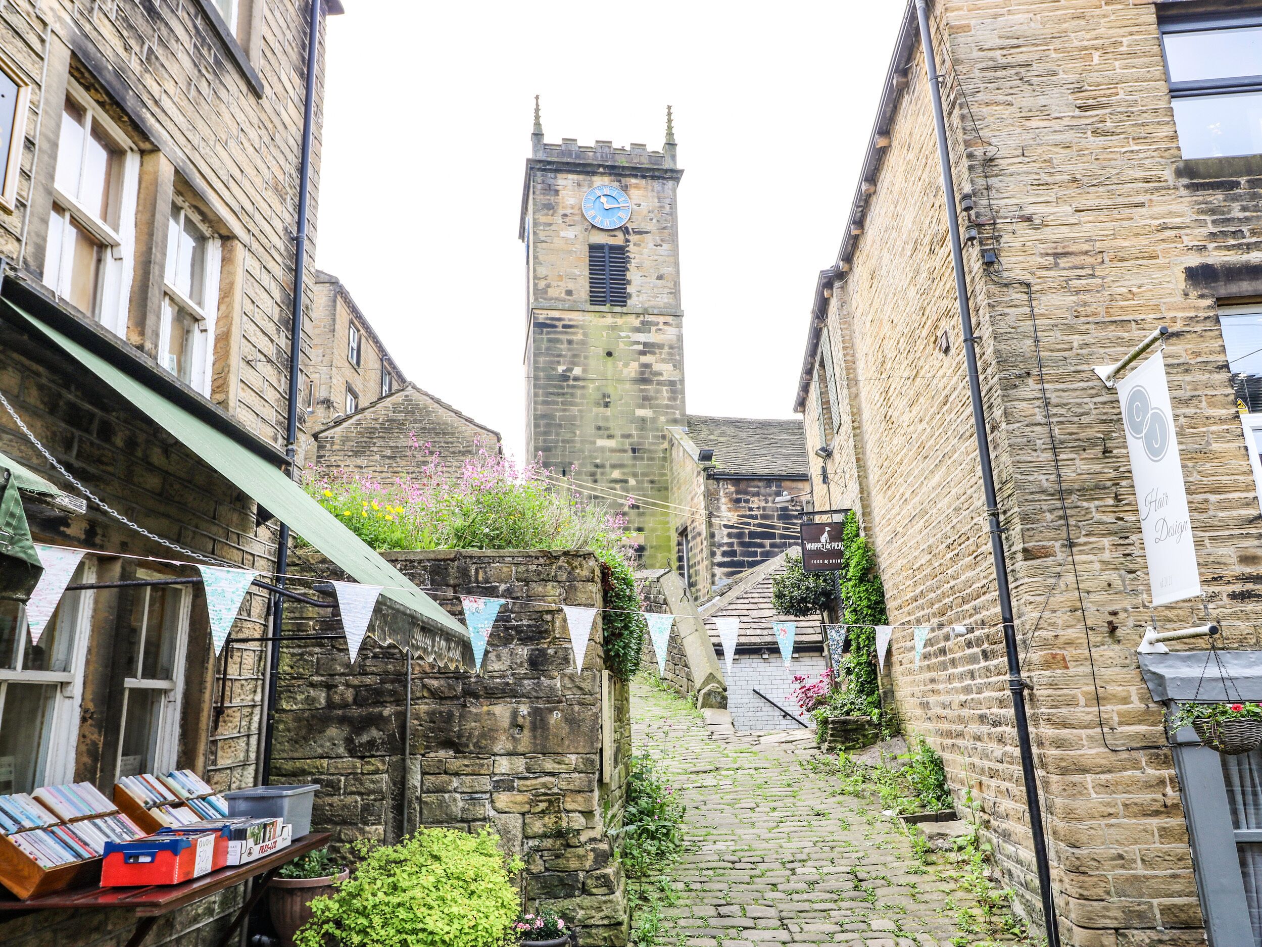 A narrow cobblestone street flanked by historic stone buildings, a clock tower, and a small shop with books and flowers.